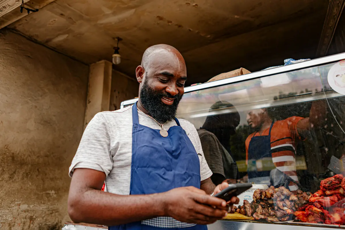 Comerciante de avental sorrindo enquanto responde mensagens no celular, atrás do balcão.