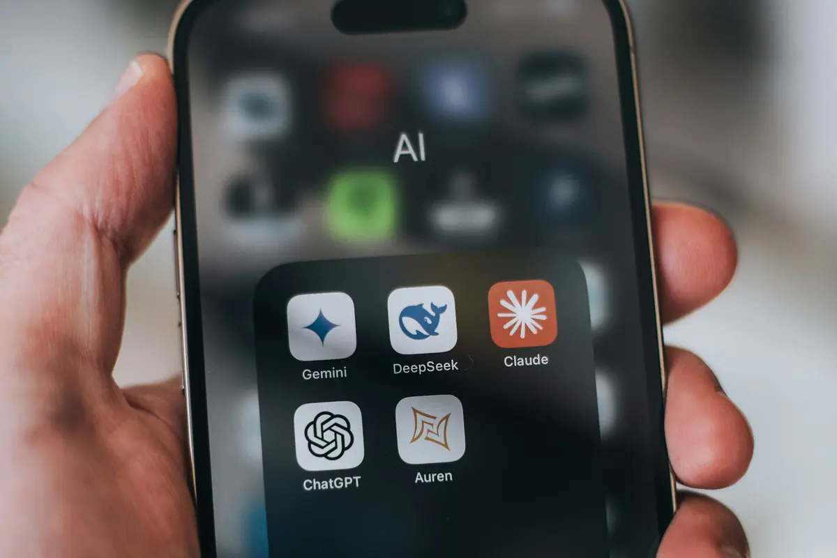 Hand holding a phone with a folder of AI apps open: Gemini, DeepSeek, Claude and ChatGPT.