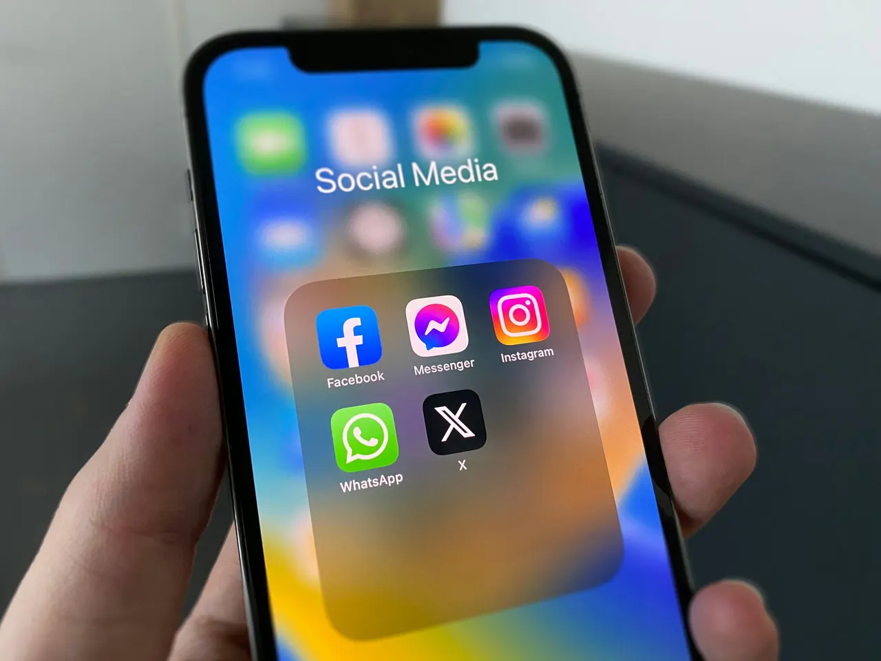 Mão segurando um celular com a pasta de redes sociais aberta: Facebook, Messenger, Instagram, WhatsApp e X.